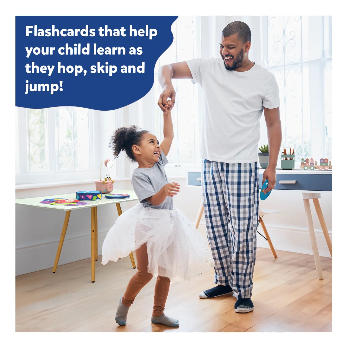 A joyful father and daughter play together in a bright, cozy room. The daughter, wearing a tutu, twirls happily while the father smiles and holds her hand. A text bubble reads, “Flashcards that help your child learn as they hop, skip and jump!” A table with colorful flashcards and a tidy play area is visible in the background.