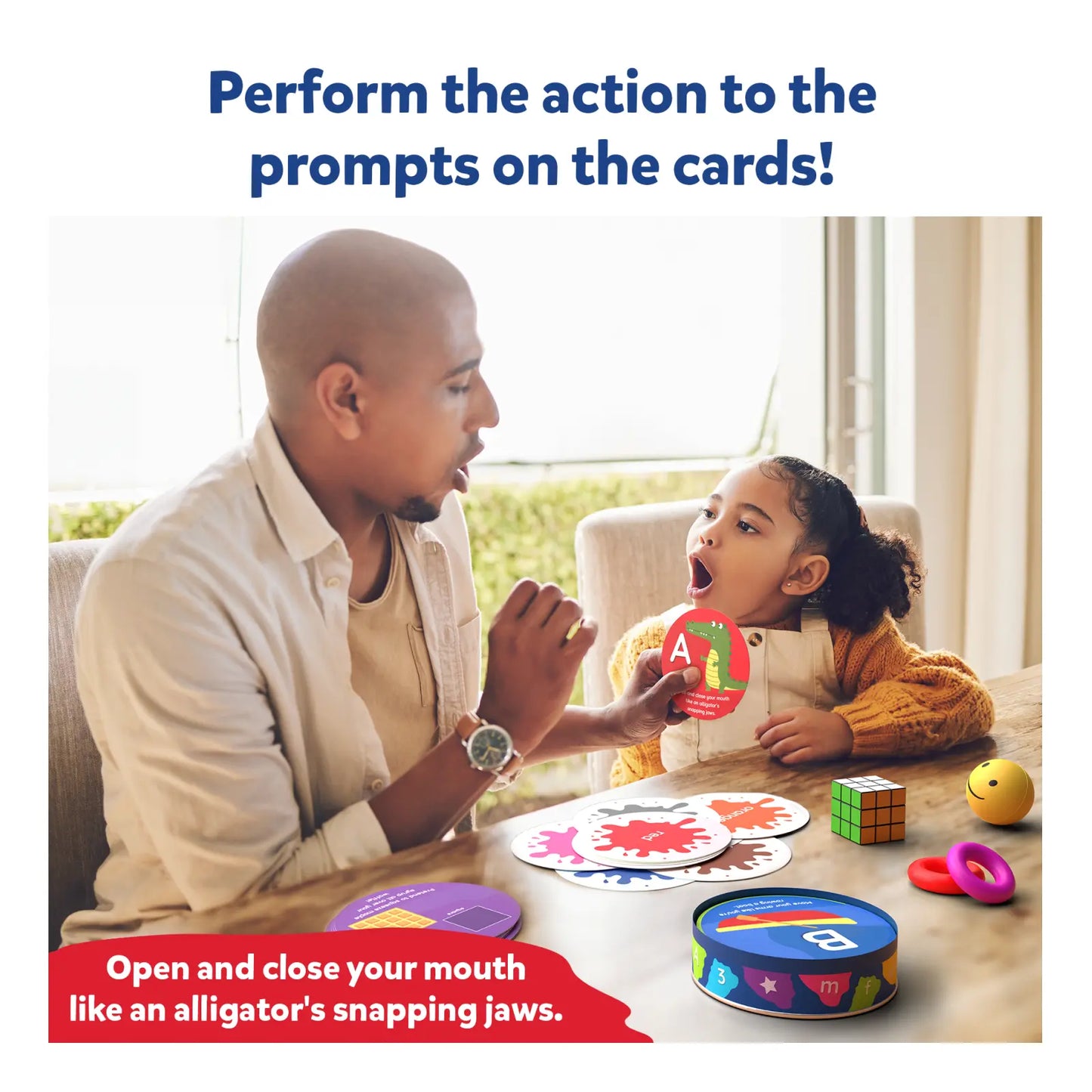 A father and daughter sit together at a table, playfully engaging with flashcards. The daughter opens her mouth wide while holding a card with the letter “A”, following the prompt, “Open and close your mouth like an alligator’s snapping jaws.” Various colorful learning tools and flashcards are spread out on the table.