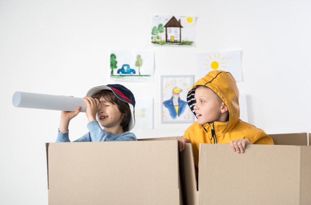 Two boys playing inside cardboard boxes and pretending they are ships. One boy is using a paper roll as a telescope.