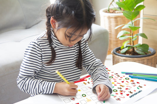 article image showing -Young girl with dark braided hair in a black-and-white striped turtleneck works on a holiday-themed activity book at a white table, holding a yellow pencil, focused, with coloured pencils nearby and a sofa and potted plants behind