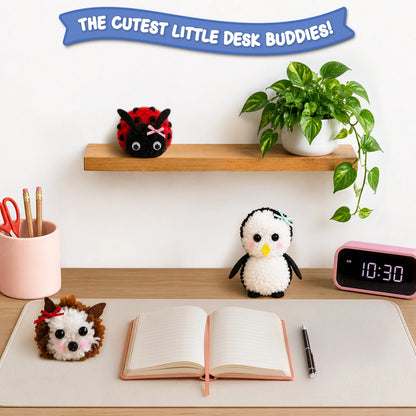 Three finished Skillmatics PomPals yarn animals—a red-and-black ladybug, a white penguin and a brown-and-white hedgehog—are displayed on a desk and shelf beside a notebook, pen, clock, stationery holder and potted plant, showing them as cute desk companions.