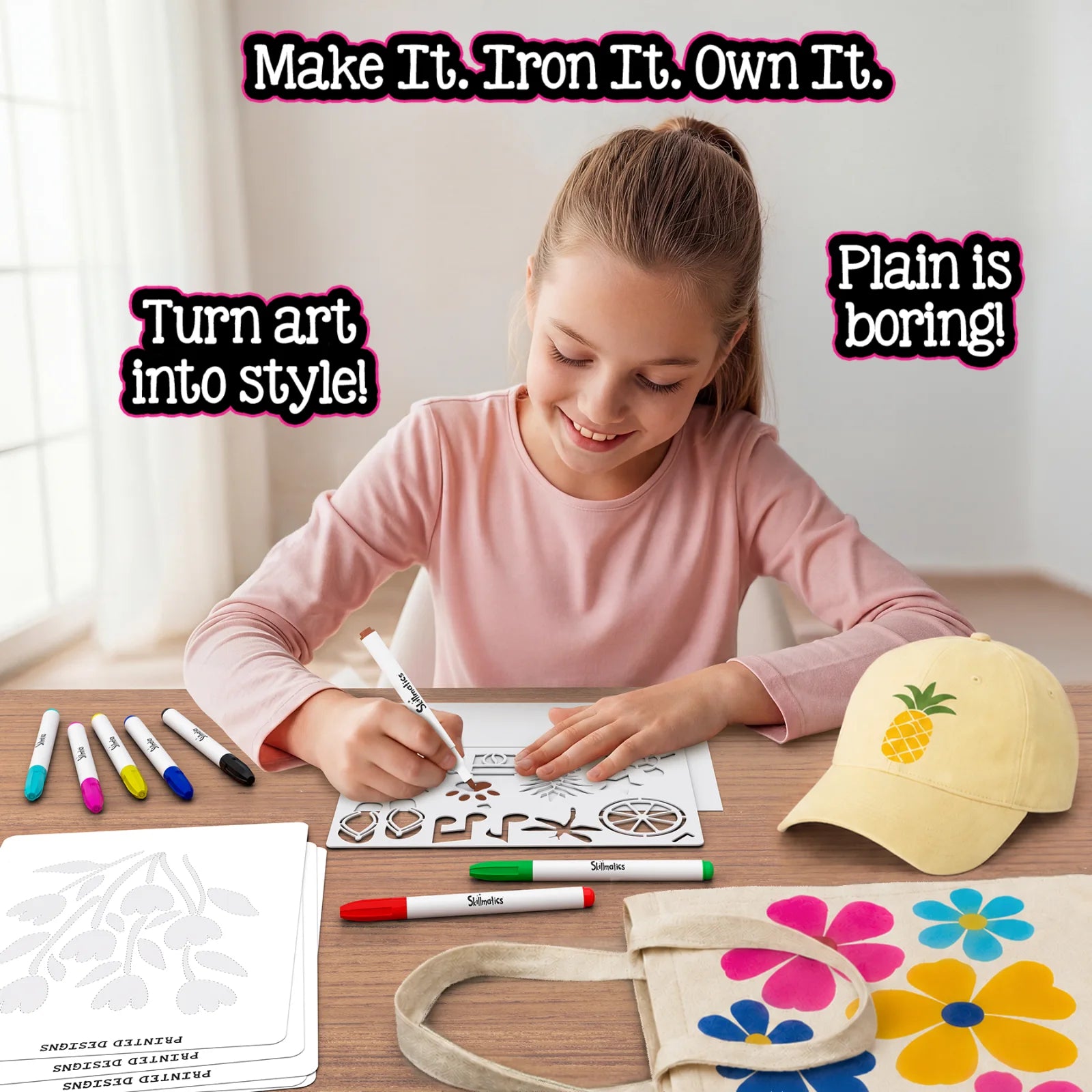 Girl with ponytail coloring a nature stencil using Skillmatics magic iron-on markers at a wooden desk. Printed design sheets, multiple colorful markers visible. Finished items shown: yellow cap with pineapple iron-on and canvas tote with colorful flower designs. Text: Make It. Iron It. Own It.