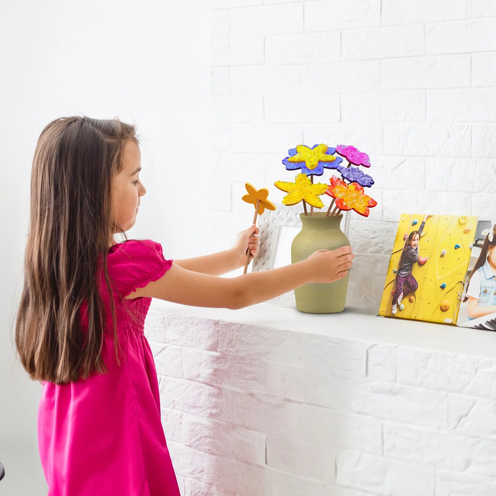Young girl in a pink dress arranging poke-in fabric flowers into a vase on a white shelf. The colorful flowers are made from the kit’s foam shapes and fabric squares.